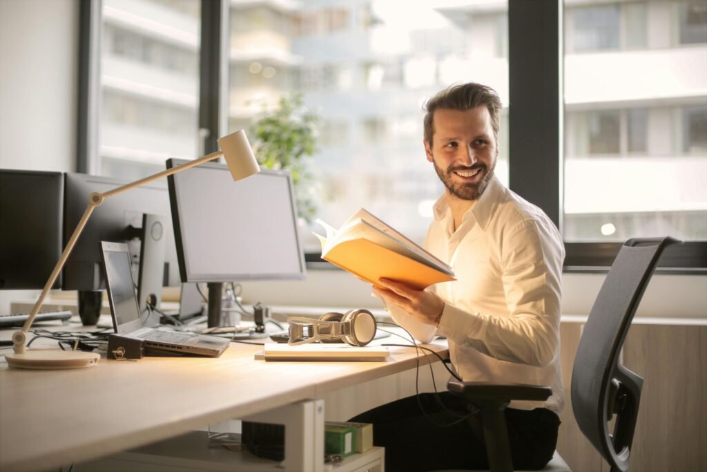 pexelsolly927022 Beawareofmecom Man in a bright office smiling and holding an open book reading about Mental Health in the Workplace with computer monitors on his desk