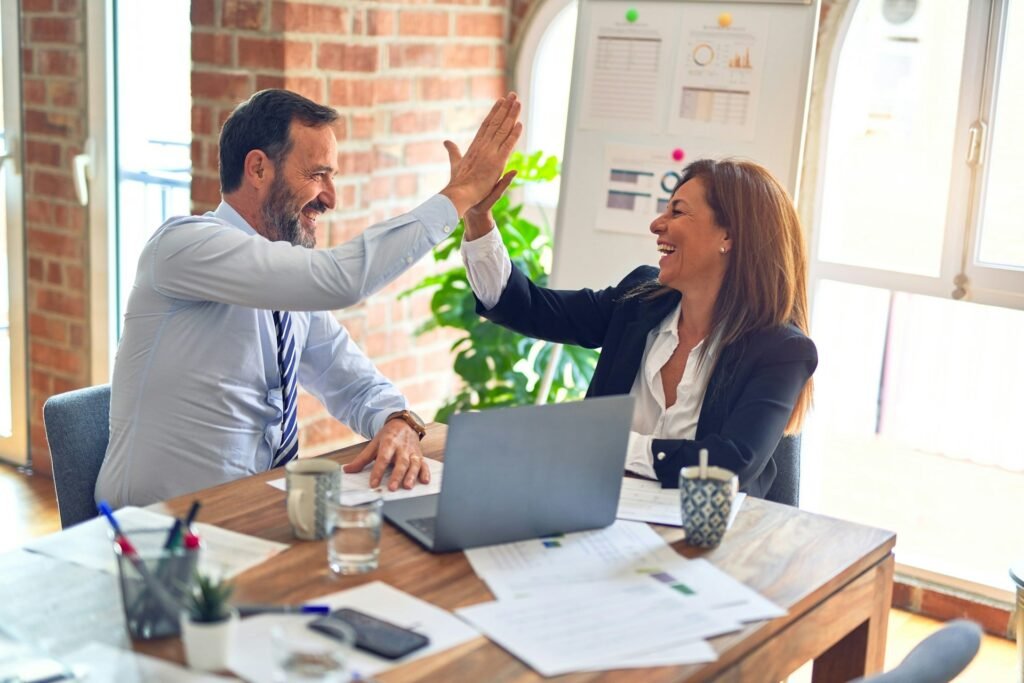Financial Inequality in Relationships How Money Impacts Mental Health and What to Do About It Beawareofmecom A professional man and woman sharing a celebratory high five in an office smiling with laptops documents and coffee mugs on the desk