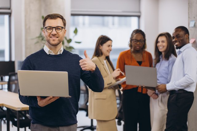 Professional working on laptop giving thumbs up with multicultural team collaborating behind in contemporary workplace setting Beawareofmecom Supportive Work Environment